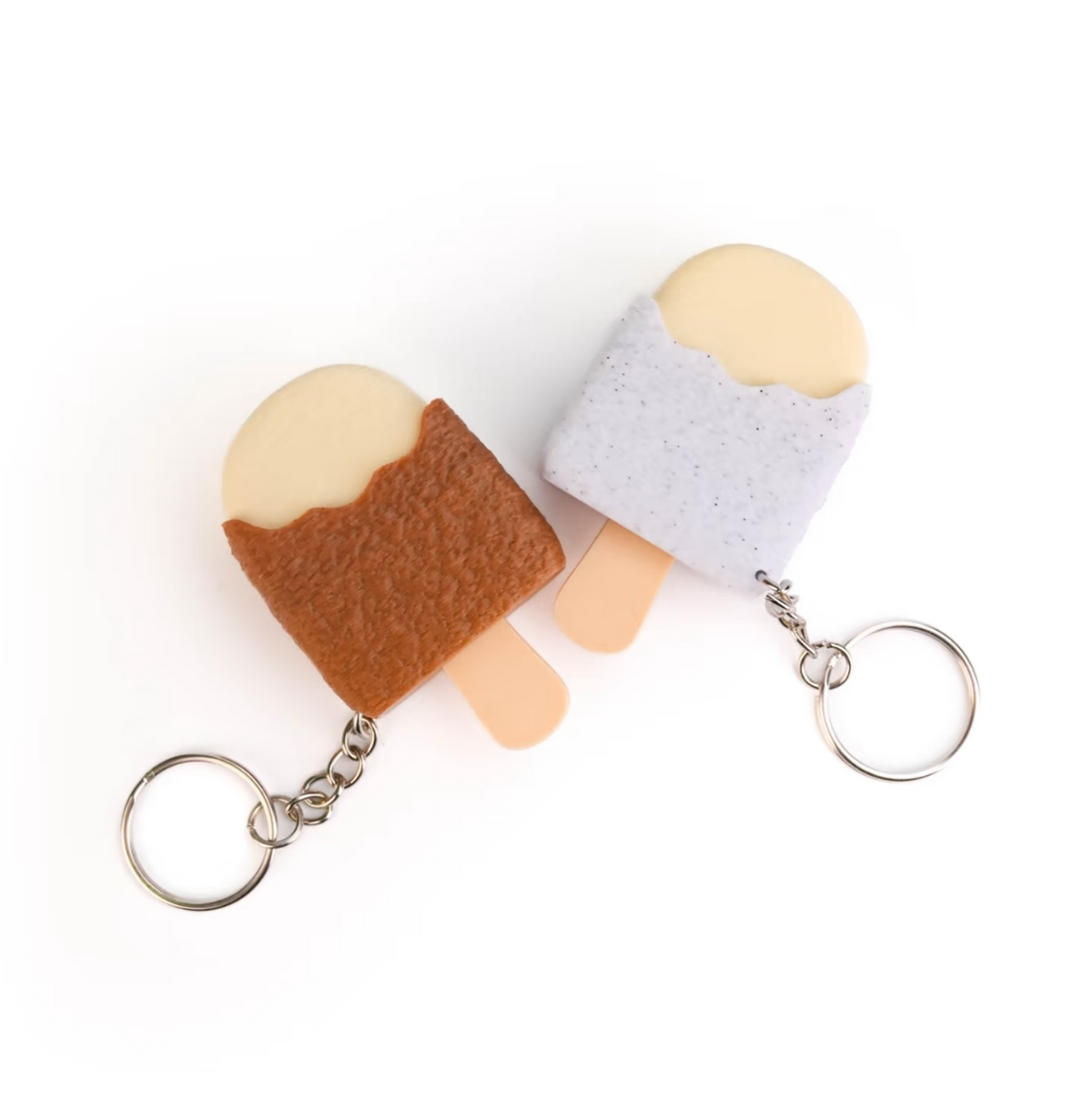Two small felt keychains shaped like popsicles on a plain white background. One has a brown body with a pale cream top and peach stick, and the other has a light lavender body with a pale cream top and peach stick.
