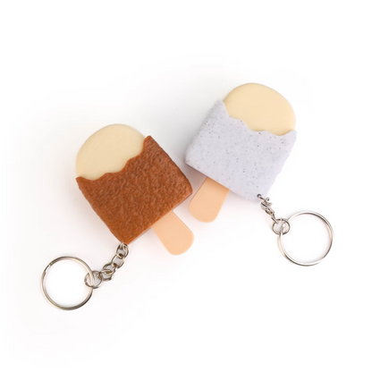 Two small felt keychains shaped like popsicles on a plain white background. One has a brown body with a pale cream top and peach stick, and the other has a light lavender body with a pale cream top and peach stick.